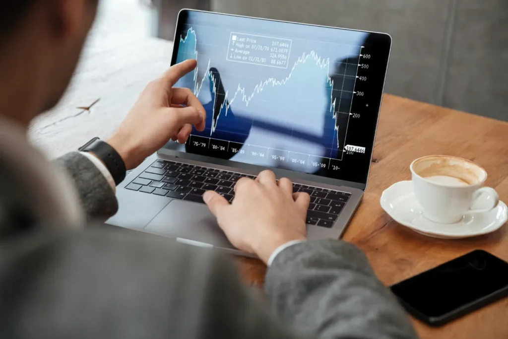 Visão de trás de um investidor analisando gráficos financeiros e dados de mercado em um notebook, com uma xícara de café ao lado.