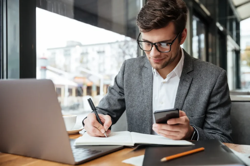 Homem de óculos, em traje social, usando notebook, celular e escrevendo em caderno, representando o perfil de um investidor focado e moderno.