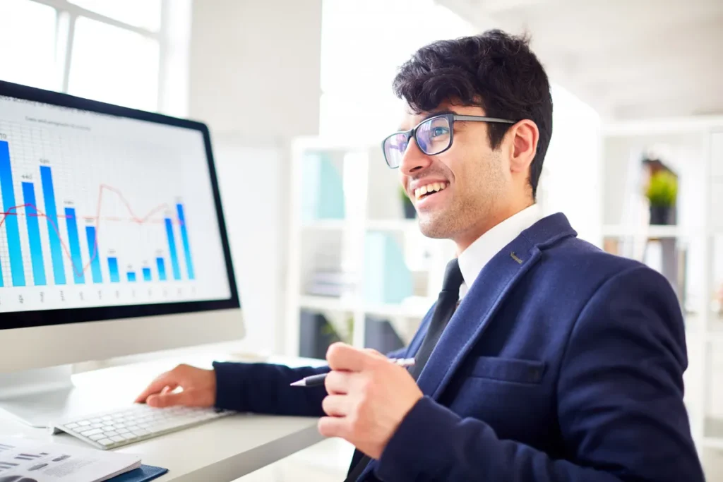 Homem sorridente com óculos, em terno, olhando para gráficos financeiros em um monitor de computador, demonstrando sucesso no perfil de investidor.