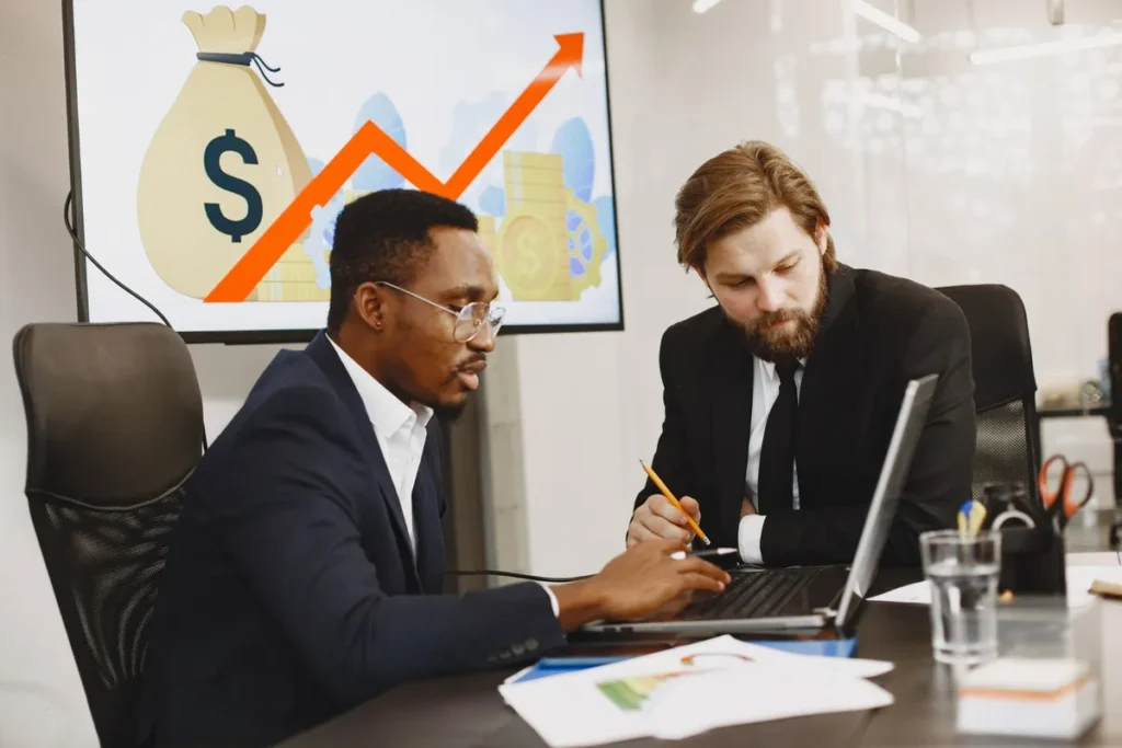 Dois homens em trajes sociais, um deles apontando para um laptop, com um gráfico de crescimento financeiro exibido em uma tela ao fundo, representando a colaboração na definição do perfil de um investidor.